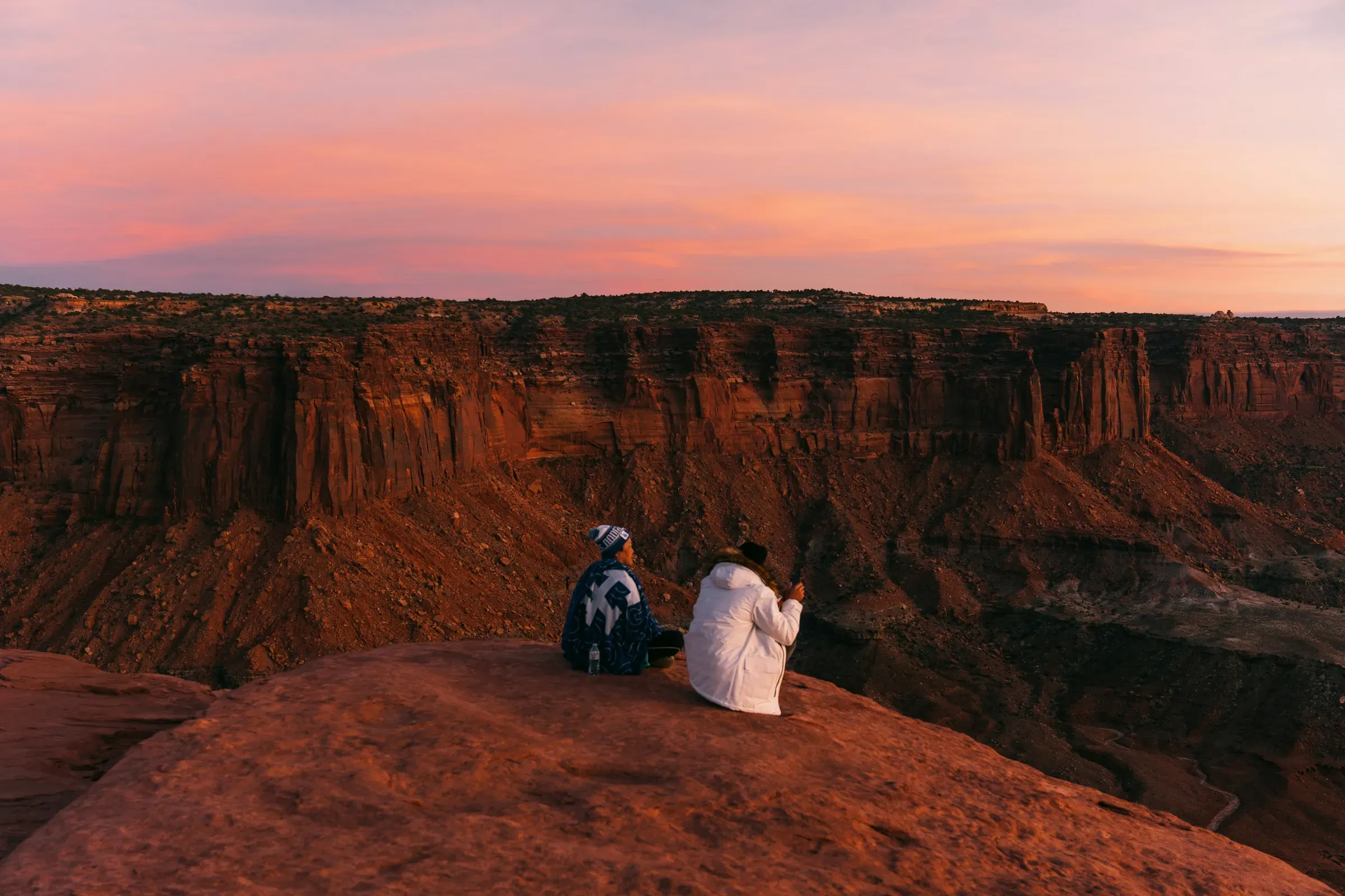 Two people sit shoulder to shoulder on a canyon rim at dusk, pink light washing over the red cliffs beyond them.